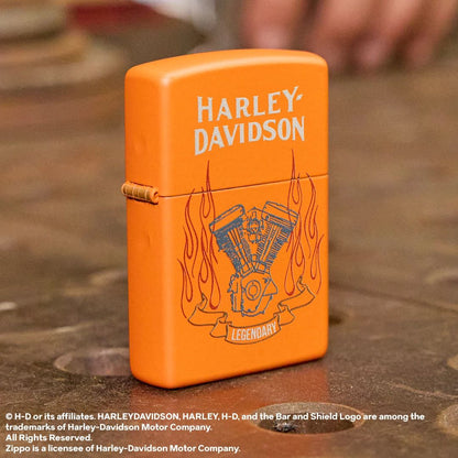 Lifestyle image of Zippo Harley-Davidson® Legendary Design Orange Matte Windproof Lighter standing at a 3/4 angle on a workshop welding table while showing the front and hinge side of the lighter. 

