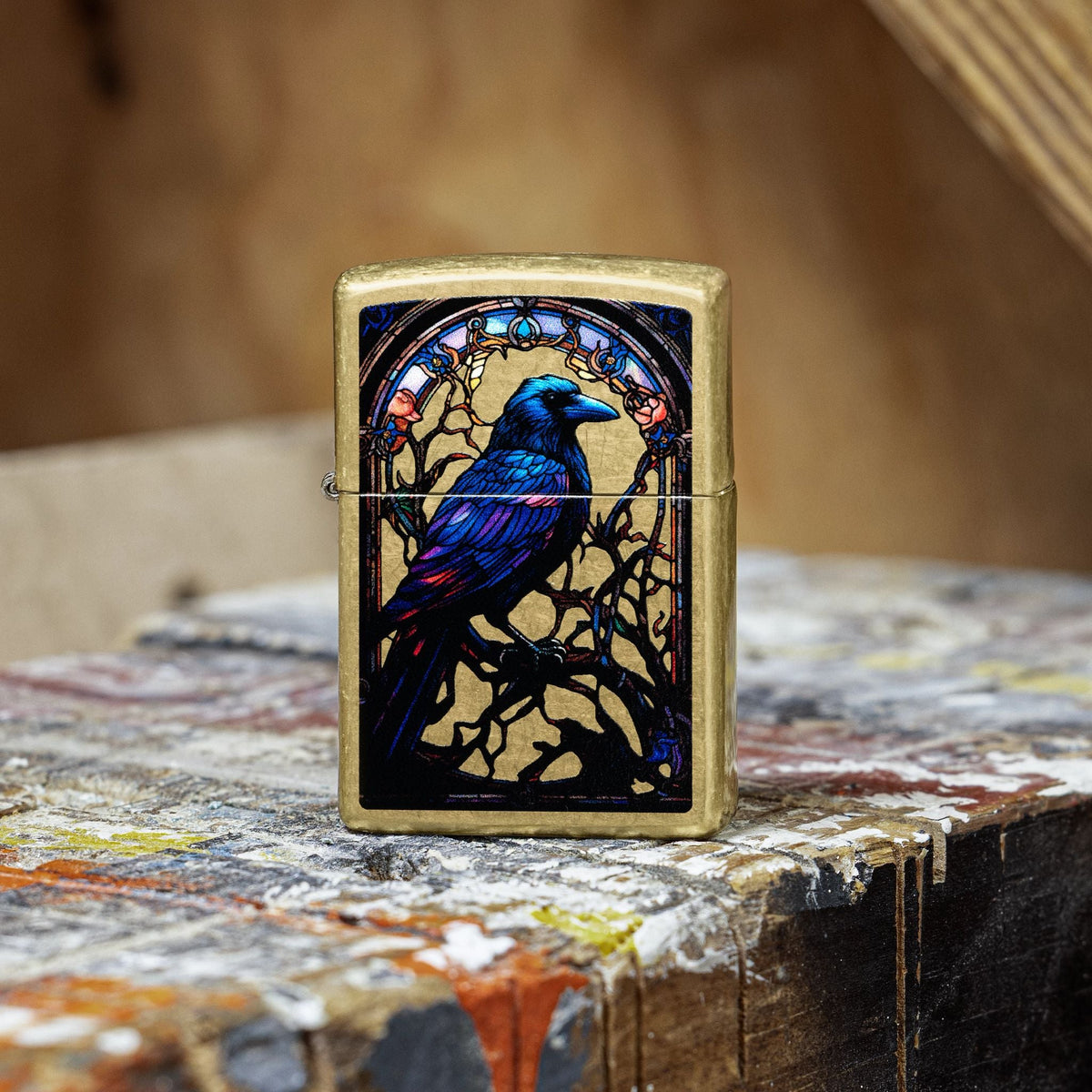 Lifestyle image of Zippo Stained Glass Raven Design Street Brass Windproof Lighter standing on a wooden box that is painted multiple colors in a graffiti style, showing the front of the lighter.  

