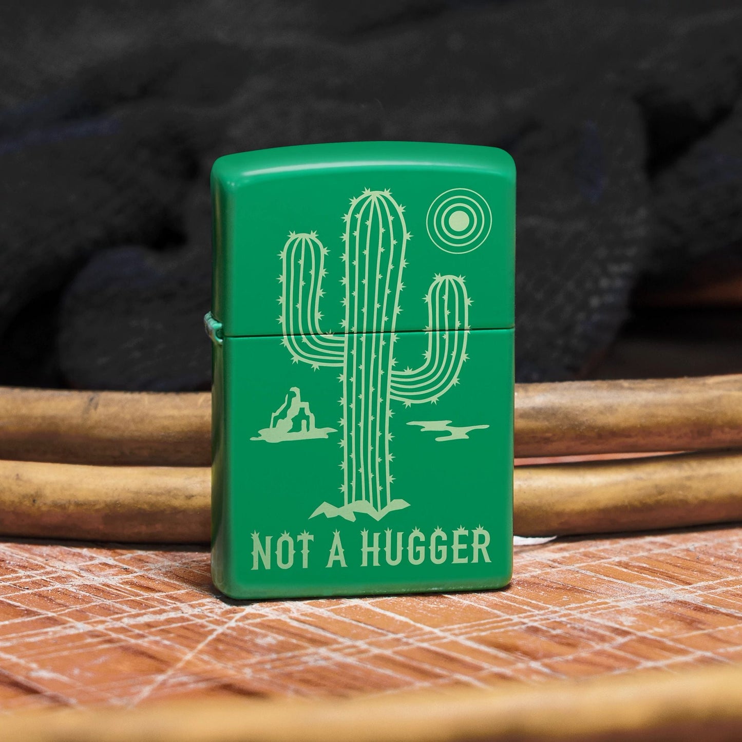 Lifestyle image of Zippo Not a Hugger Design Grass Green Matte Windproof Lighter standing on a wooden surface in front of a few power cords, showing the front of the lighter. 

